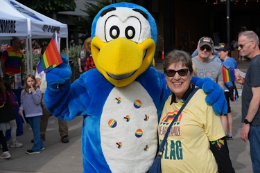 a blue bird mascot with rainbow stickers on the belly hugs an older woman